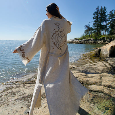 A person standing by the water wearing a beige organic linen kimono with hand-block printed snake symbol on the back.